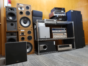 An organized display of vintage audio equipment, including large wooden and black speakers, amplifiers, receivers, and subwoofers arranged on metal shelving against a wood-paneled wall. The setup highlights classic home audio systems and components, showcasing both functionality and nostalgia for older audio technology.