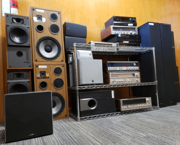 An organized display of vintage audio equipment, including large wooden and black speakers, amplifiers, receivers, and subwoofers arranged on metal shelving against a wood-paneled wall. The setup highlights classic home audio systems and components, showcasing both functionality and nostalgia for older audio technology.